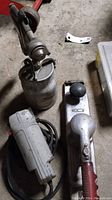 Air belt sander, Husky air paint sprayer, and electrical drywall saw on concrete floor with some garage dust and a small miscellaneous metal piece nearby.