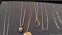 Full view of four gold-tone necklaces laid out on a dark surface, showing different chain styles and pendants including a lion head and a leaf.