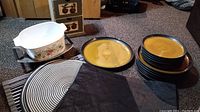 View of black placemats, cookie cooling racks, CorningWare dish, plates and platter arranged on floor.