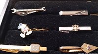 Photo showing various men's tie clips including a wrench-shaped BLACKHAWK clip, a crocodile design clip, and a cartoon character clip among others.