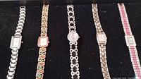 Five watches displayed on black fabric background showing variety of metal link bands and designs, including rhinestone embellishments, rectangular and round faces.