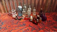 Front view of all 20 old bottles arranged on a red patterned fabric, showing variety in bottle shapes and colors including a tall blue Ball jar and several clear and amber bottles.
