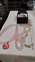 Shot of two pink necklaces laid flat: one mesh wire pink cord necklace with heart-shaped pink stone pendant and one pink bead necklace, next to a black box containing the crystal paperweight.