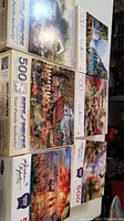 Top view of six 500-piece puzzle boxes with colorful scenic images including a covered bridge, meadow farms, deer in an autumn forest, trick or treat Halloween scene, and Rocky Mountain campsite.