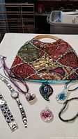 A small, multicolored patchwork fabric handbag with bamboo handles sits behind two silver-tone watches, four glass pendant necklaces laid out on a white cloth, and a beaded bracelet.