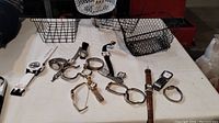 Wide photo showing 17 watches laid flat on white surface along with four black wire baskets.
