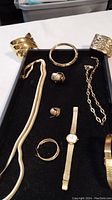 Overview of woman's gold-tone jewelry including necklaces, rings, bracelets, earrings, and watches on black display tray.