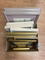 Photo showing tops of eleven framed prints stacked in a box, showing variety of frame colors and thicknesses.