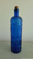 Front view of the tall cobalt blue glass bottle showing grapevine embossed pattern and cork stopper