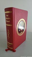 Side view of The Catholic Missal showing spine title and red cover with gold embossed details and red ribbon bookmarks.