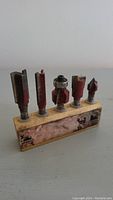Five wood router bits of various shapes and sizes stored in a worn wooden holder, placed on a gray surface
