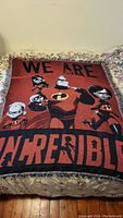 Photo showing The Incredibles tapestry blanket fully spread on a bed, depicting main characters and the text 'WE ARE INCREDIBLE'.