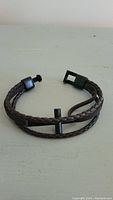 Photo showing the coiled brown braided leather bracelet with black metal cross and clasp, placed on a flat light-colored surface.