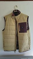 Full view of beige quilted Vans vest hung on hanger showing front and chest pocket with dark brown fabric and zipper.