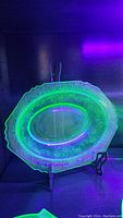 Oval uranium glass bowl glowing bright green under UV light, showing floral etching and scalloped edges on display stand.