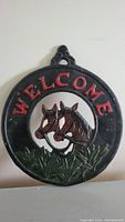 Front view of black cast iron welcome sign with red lettering and two horses' heads with bridles, with green grass detail at the bottom.