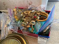 Photo showing a box filled with assorted costume jewelry including necklaces and bracelets, mostly in plastic bags. Colors and materials vary, including wooden beads and gold-tone metals.