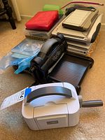 Photo showing Big Shot die cutter in black, Stampin Up die cutter in white, guillotine paper cutter, and various plastic storage boxes with crafting tools and dies on carpet floor.