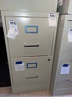 Front view of beige metal 2 drawer filing cabinet with locked upper drawer and label holders taped with blue tape.
