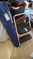 Metal folding step stool with two steps standing next to blue carrying bag on floor.