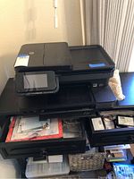 HP Photosmart 7520 printer with touchscreen control panel and paper tray visible, showing the front and side views of the device on a black storage cabinet.