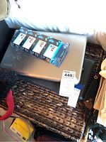 Silver Toshiba laptop with visible wear, placed on wicker surface alongside boxed HP 564 ink cartridges.