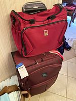 Photo of two red luggage pieces stacked, showing upright suitcase with wheels and collapsible handle, with duffle bag on top showing front zippered pocket and carrying handles.