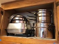 Image of assorted stainless steel pots and pans in a wooden cabinet showing stacked cookware including pots and pans with black handles and some lids.