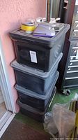 Stack of three large black plastic tubs with lids stored outside near pink wall and door. Tubs appear full and sealed.