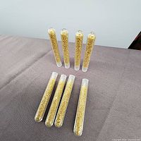 Photo showing eight clear test tubes filled with small 24k gold flecks arranged on a gray surface.