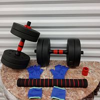 Photo showing adjustable dumbbells, blue fingerless gloves, red collars, and foam-covered barbell connector bar on a marble surface.