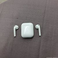Top view of the Apple AirPods with charging case on a purple fabric surface, showing one left earbud, one right earbud, and the charging case in the center.