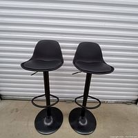 Two black bar stools with curved backrests, adjustable height lever, metal base with round footrest on concrete floor in front of a metal roll-up door.