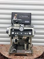 Front view of espresso machine with integrated grinder, portafilter attached, milk pitcher and tamper on drip tray with original box behind