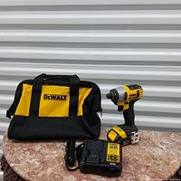 Photo of DeWalt impact drill with battery attached, battery charger, and branded carrying bag on a round marble table surface.