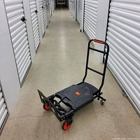 Image shows a new folding hand truck with black metal frame and orange wheels placed in a storage hallway.