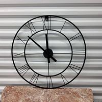 Front view of the metal wall clock showing open design and Roman numerals with black clock hands.