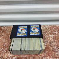Black storage box filled with a large collection of standard-sized Pokemon cards showing the backs of cards with the classic Pokemon blue and Pokeball design.