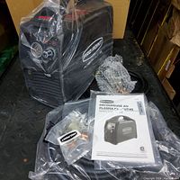 Main plasma cutter unit in black steel housing with pressure gauge and control dial, torch and ground cable wrapped in bubble wrap, consumables kit and manual on metal surface