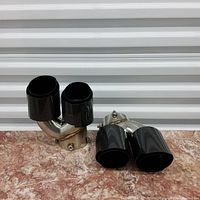 Photo of two stainless steel exhaust tips with carbon fiber dual outlets on marble surface, front view.