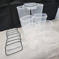 Eight clear plastic airtight food storage containers with white lids and black sealing gaskets arranged on a soft white surface against a black backdrop.