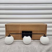 Photo of a 3-globe bathroom light fixture with white glass globes on a black metal horizontal bar, placed in front of a cardboard box on a gray soft surface.