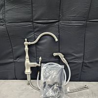 Brushed nickel single handle kitchen faucet with curved spout and side spray, placed against a black fabric background. Includes flexible braided hoses and mounting hardware packed in a plastic bag.