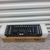 Photo of DMX 512 stage light controller in black with 16 faders and multiple buttons, resting on foam padding atop a cardboard box marked 'DMX512 controller'.