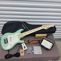 Electric guitar kit with mint green guitar, black Donner carrying bag, black and white amplifier, strap, capo, tuner, picks, and cables arranged on a table.