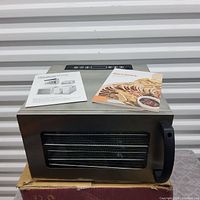 Front view of stainless steel food dehydrator with digital control panel on top, glass door, and black handle. Recipe booklet and instruction manual placed on top.
