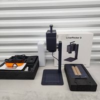Overview of LaserPecker 3 unit on stand, base plate, packaging box, foam trays with safety goggles, cables and roller attachments