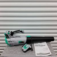 Photo of Litheli 20V cordless leaf blower with attached 20V battery, battery charger, and user manual, all placed on a lace-patterned surface.