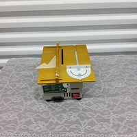 Mini table saw placed on lace-covered surface with front and top view showing yellow table surface, adjustable fence, angle gauge, motor housing, and power switch.