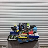 Photo showing a mixed lot of various boxed electronic and computer-related items arranged on a small table in front of a corrugated metal background.